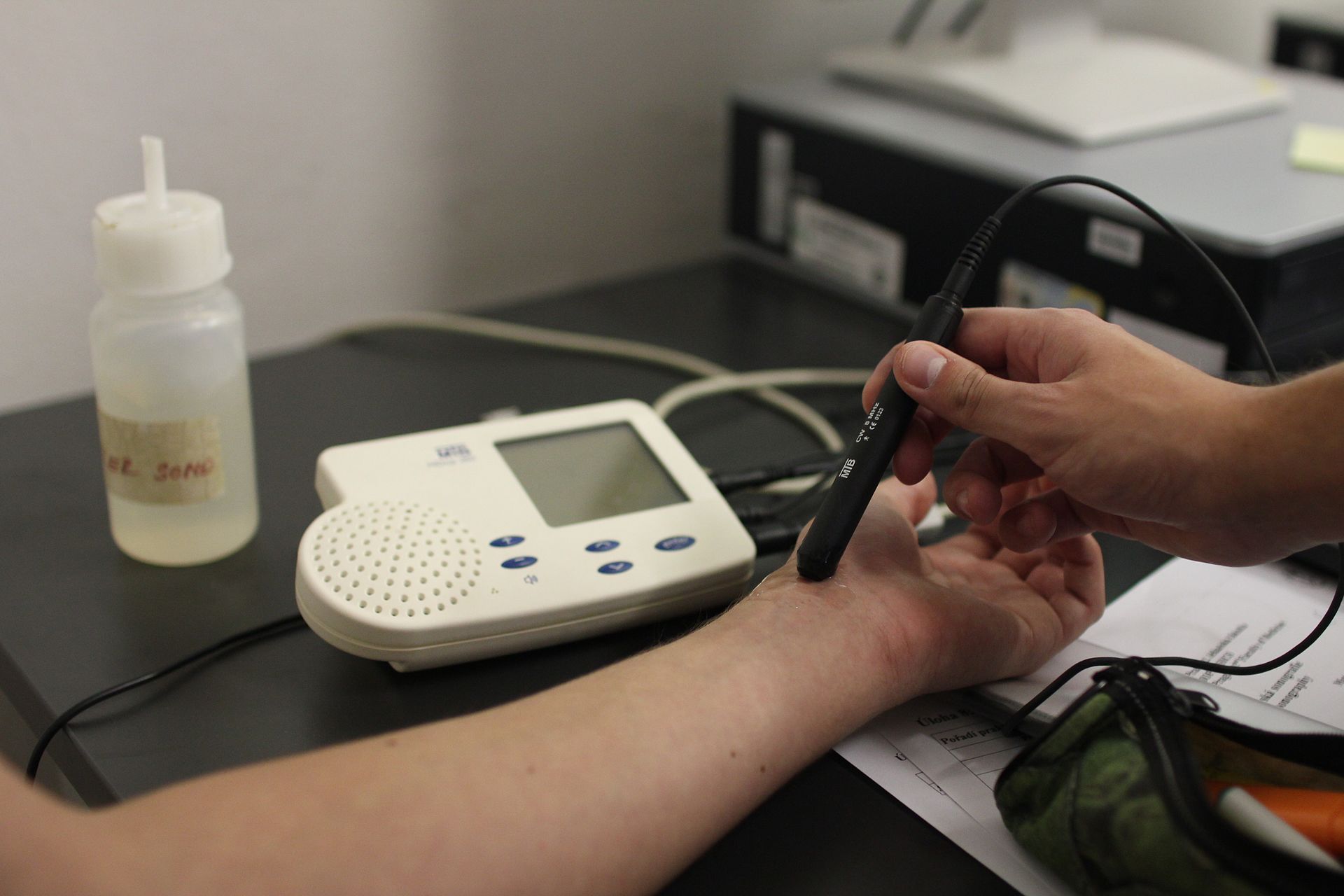 A hand holding a pencil continuous-wave Doppler probe against a wrist, beside a small Doppler unit.