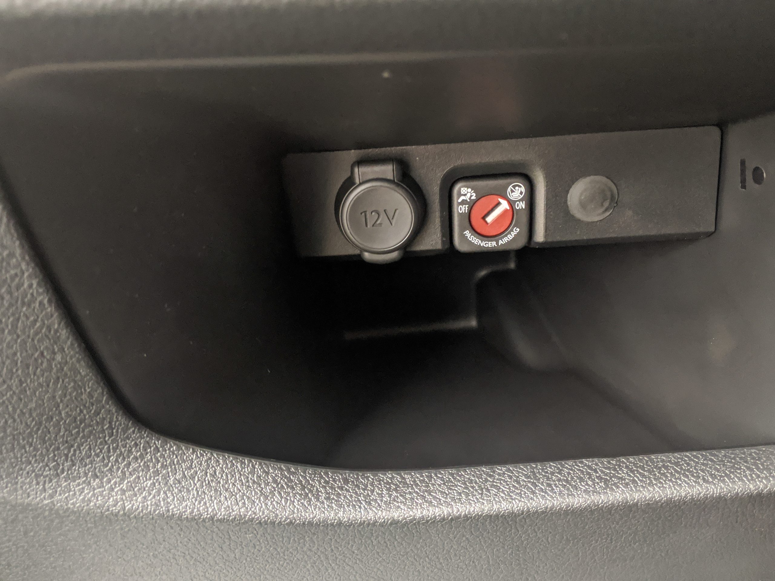 A capped 12V accessory socket in a car dash, beside a red passenger-airbag switch