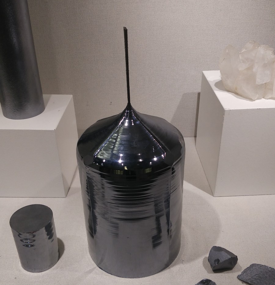 A cylindrical monocrystalline silicon ingot with its seed crystal spike on museum display beside chunks of polysilicon