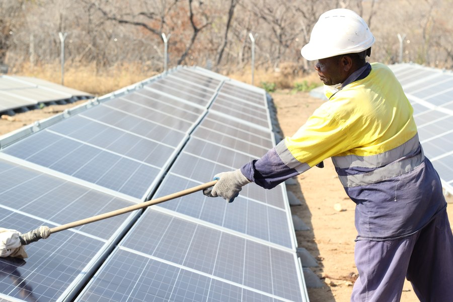 A worker in high-visibility clothing washing the surface of a ground-mounted solar array with a long-handled mop in a dry dusty landscape