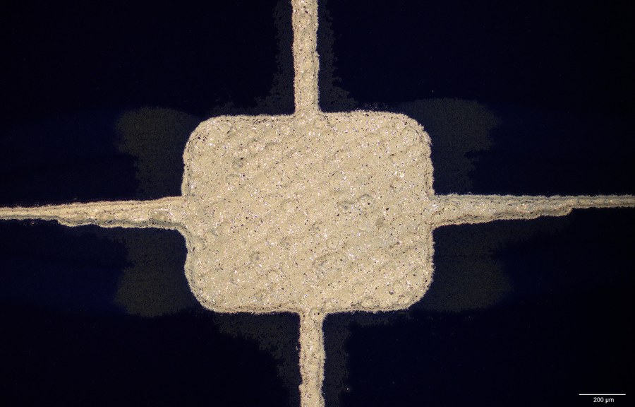 Microscope image of a screen printed silver solder pad on a solar cell where two fingers cross, showing the granular sintered paste against the dark cell surface