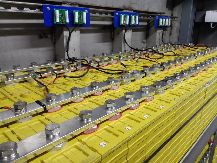 A bank of prismatic lithium iron phosphate cells joined across their terminals by flat aluminium busbars, with numbered cell tops and small balancer boards mounted on a rail above them