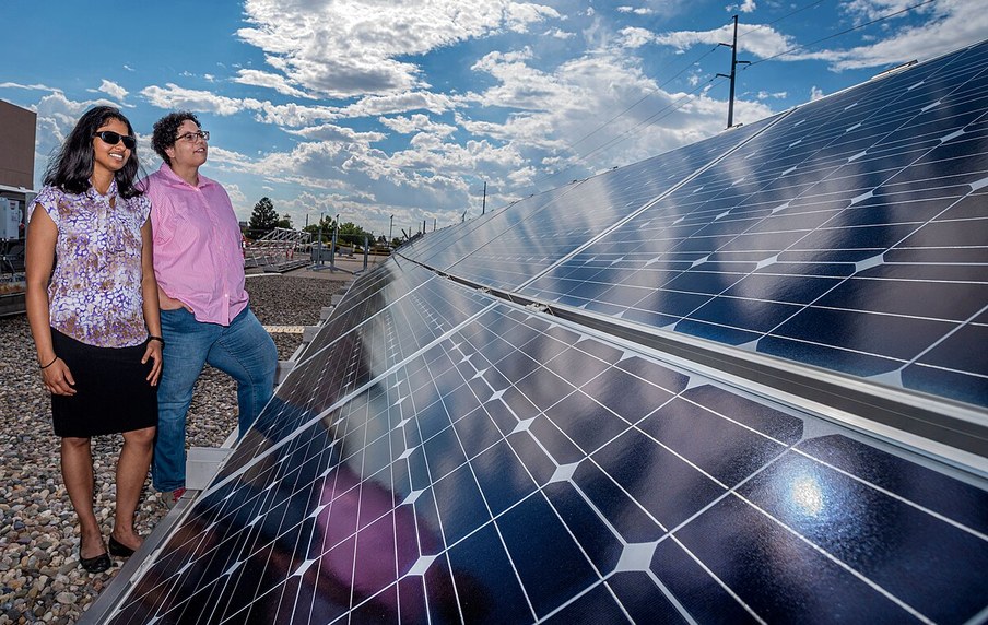 Two researchers standing beside a rack of monocrystalline photovoltaic modules on an outdoor test field under a partly cloudy sky