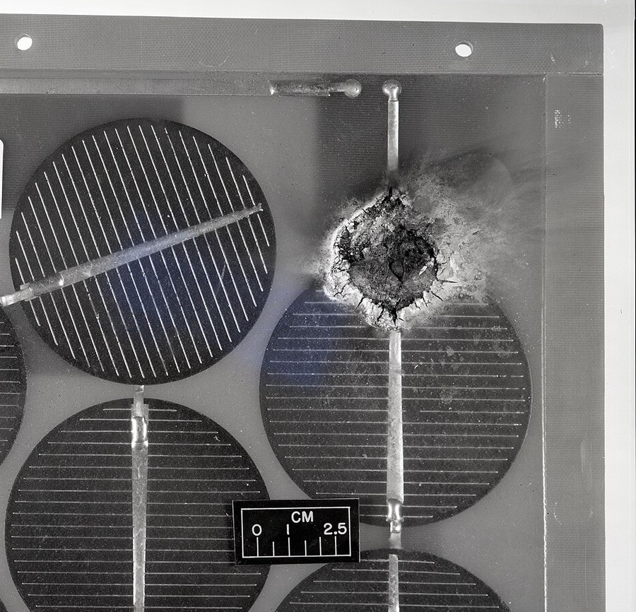 Close-up of a solar module interior showing a charred crater burned through the encapsulant beside a metal interconnect ribbon, with round cells and a centimetre scale bar