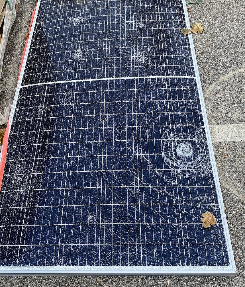 A photovoltaic module lying on tarmac with its tempered glass shattered across the whole surface, radiating cracks around a single impact point, frame and cells still intact underneath